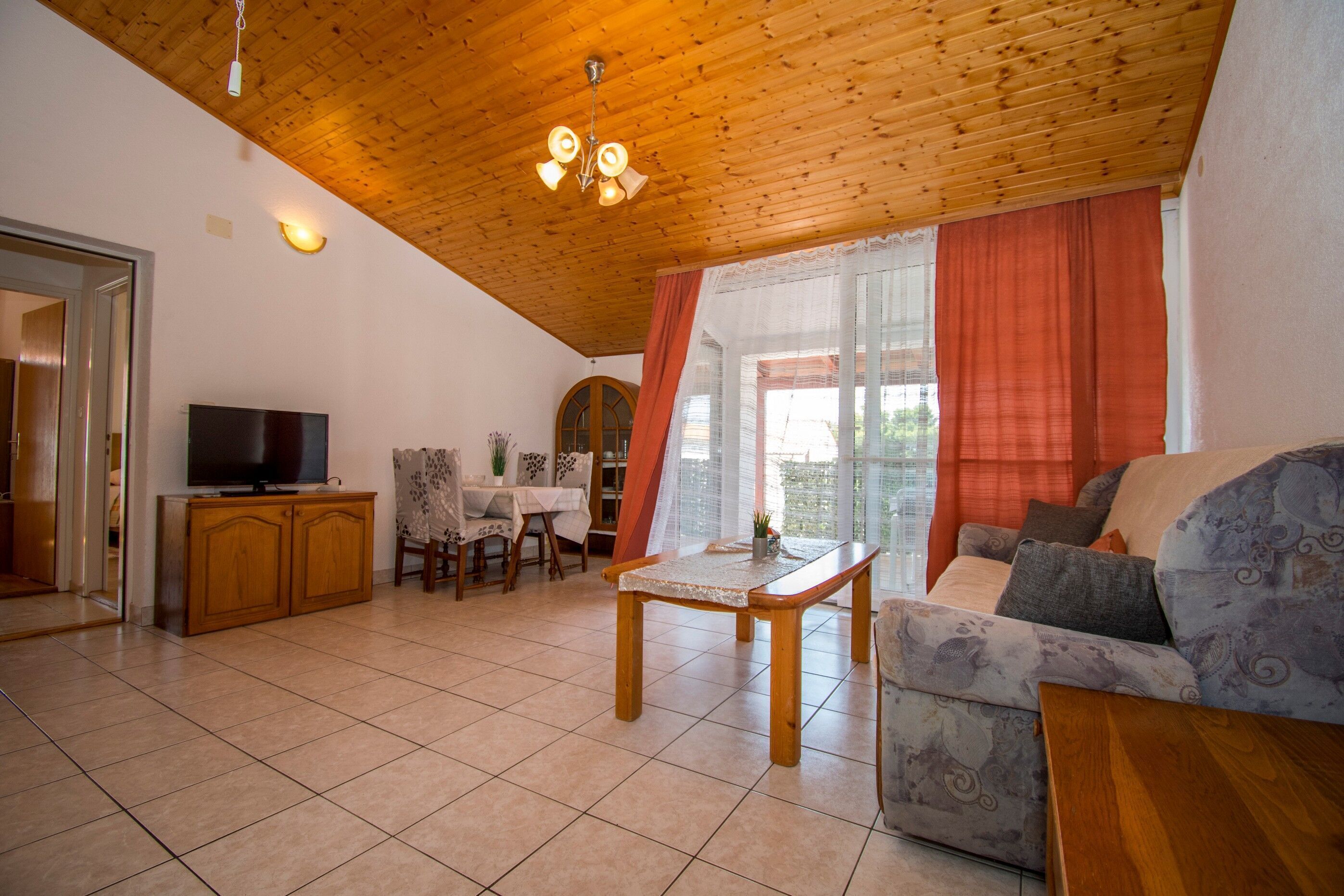 Ein Wohnzimmer mit Holzboden, einem Fernseher, einem Sofa, einem Tisch und einem Fenster mit Vorhängen.