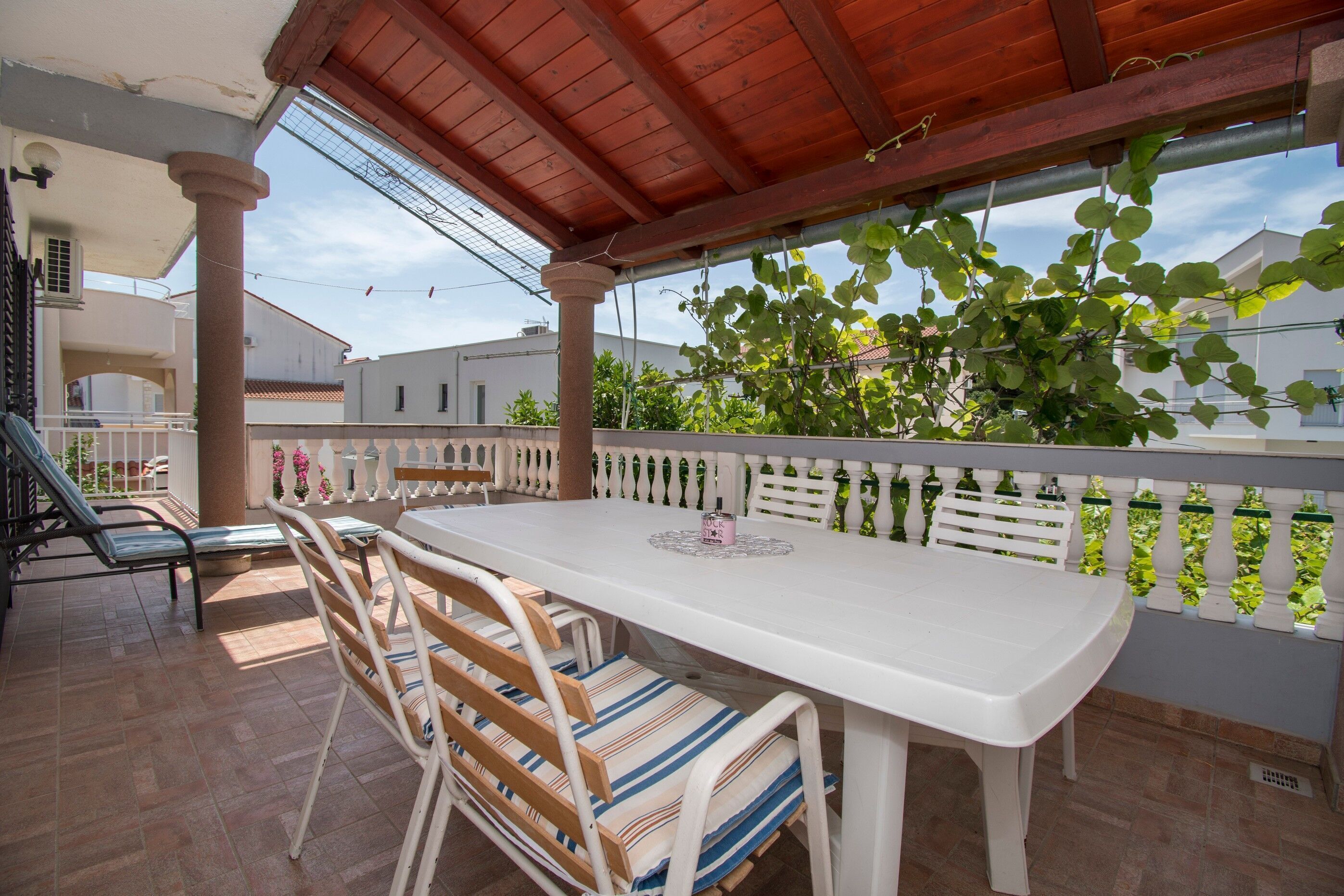 A covered patio with a white table and chairs, a wooden bench, and a view of a white fence and greenery.