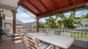 A covered patio with a white table and chairs, a wooden bench, and a view of a white fence and greenery. - Apartments Neve (Vodice)