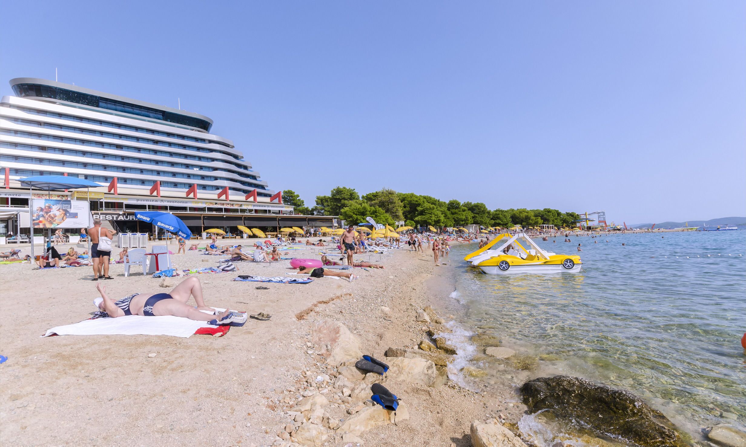 En strand med et stort hotel i baggrunden, liggestole, parasoller og en båd på vandet.
