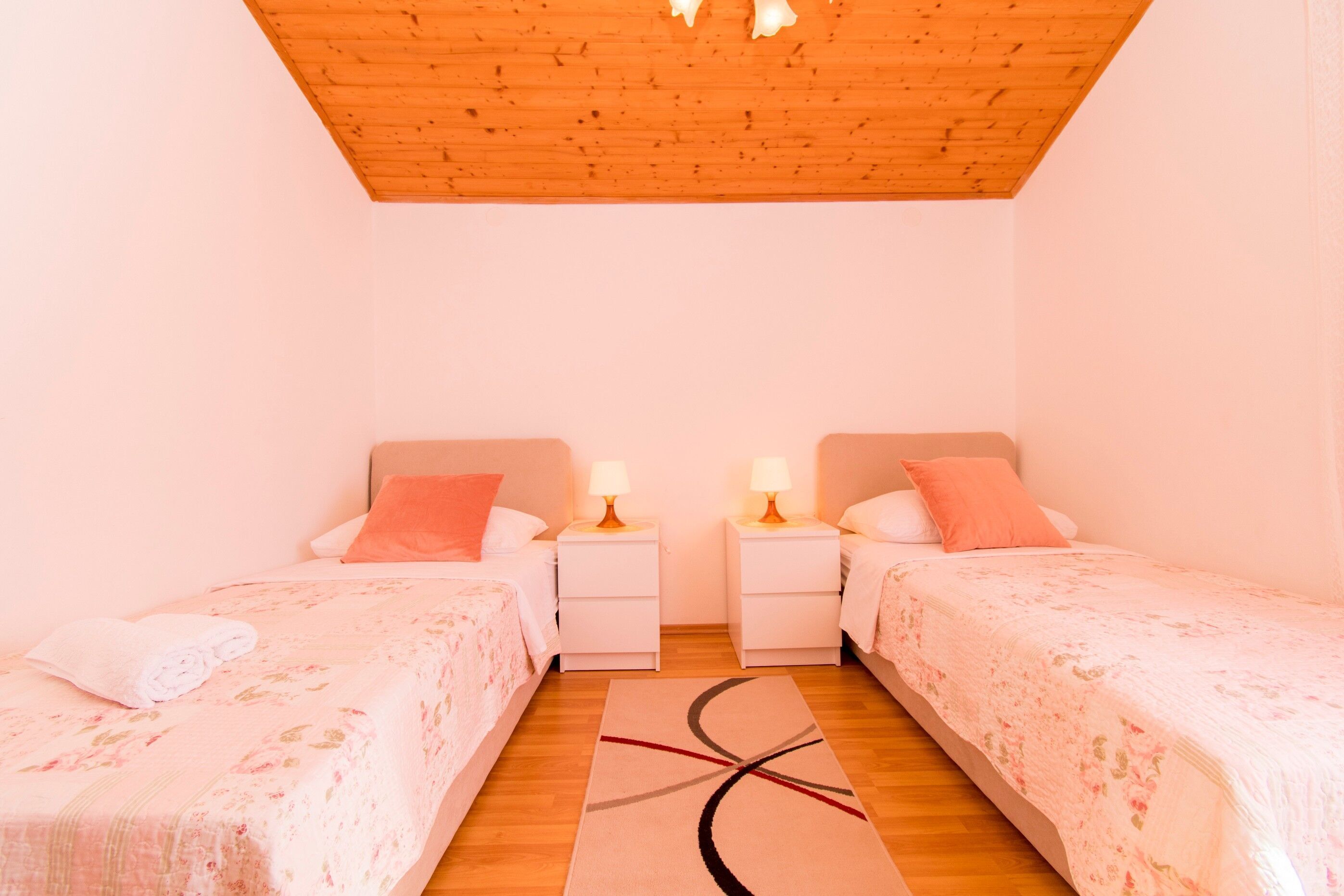 A bedroom with two single beds, nightstands with lamps, a wooden ceiling, and a floor rug.