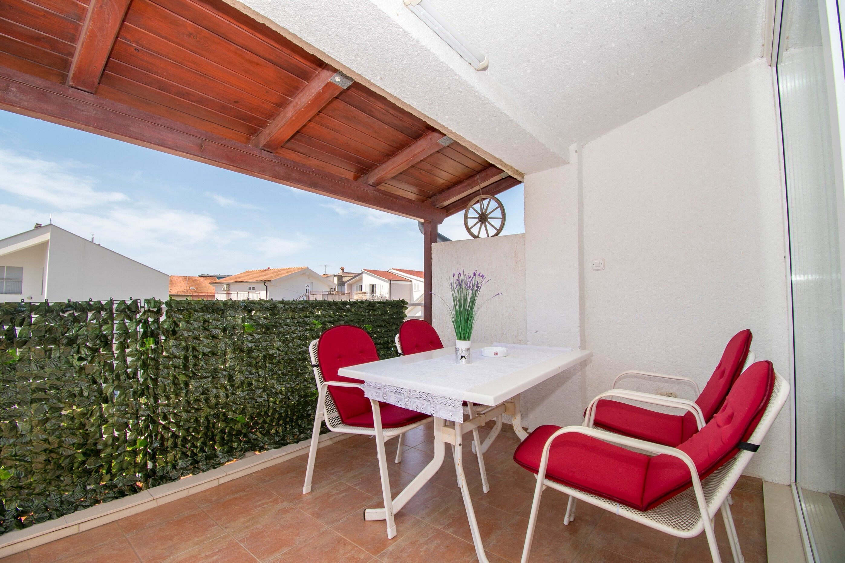 A patio with a white table and red chairs, a wooden ceiling, and a green hedge in the background.