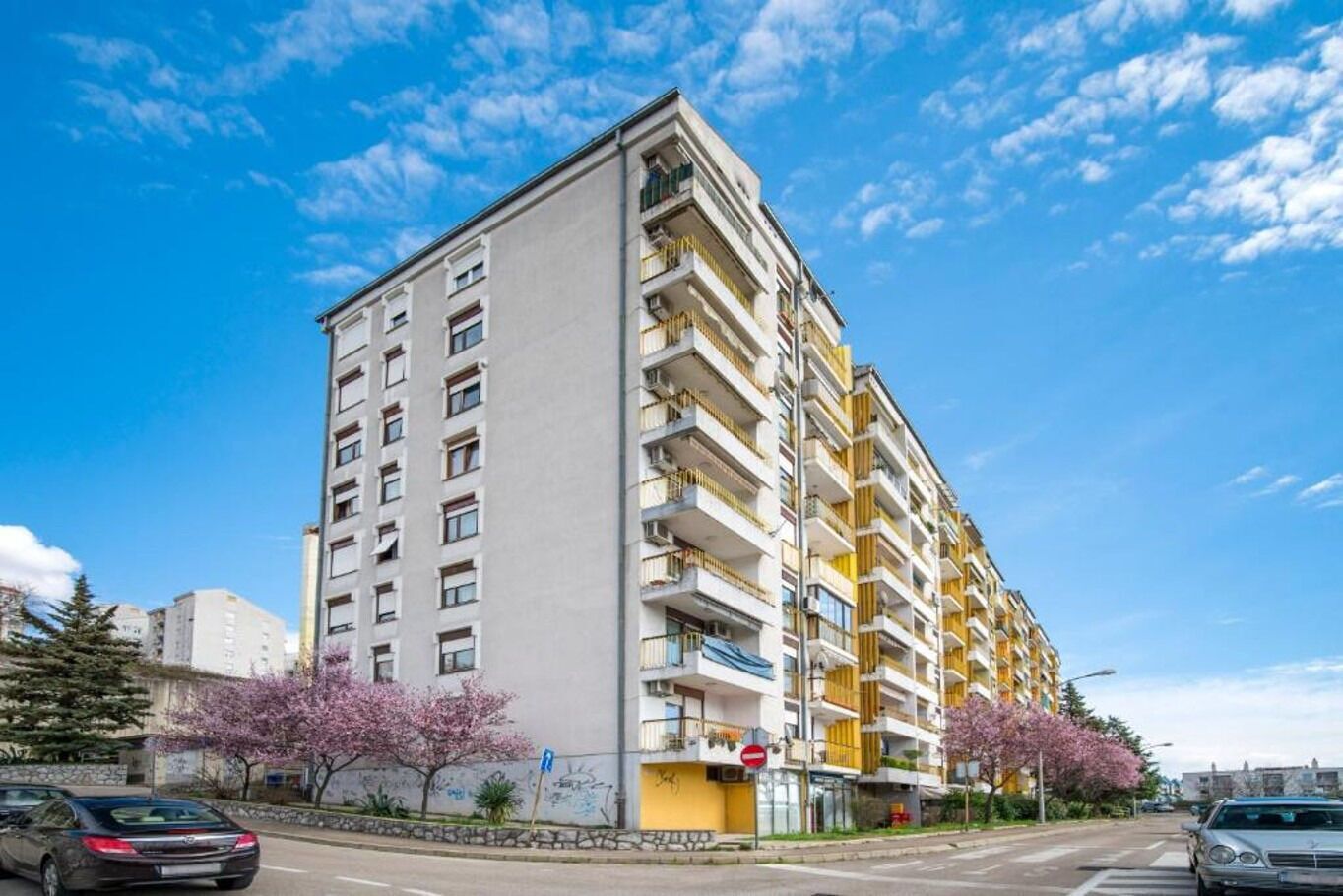 Um edifício residencial com vários andares, varandas, uma parede de pedra e cerejeiras.