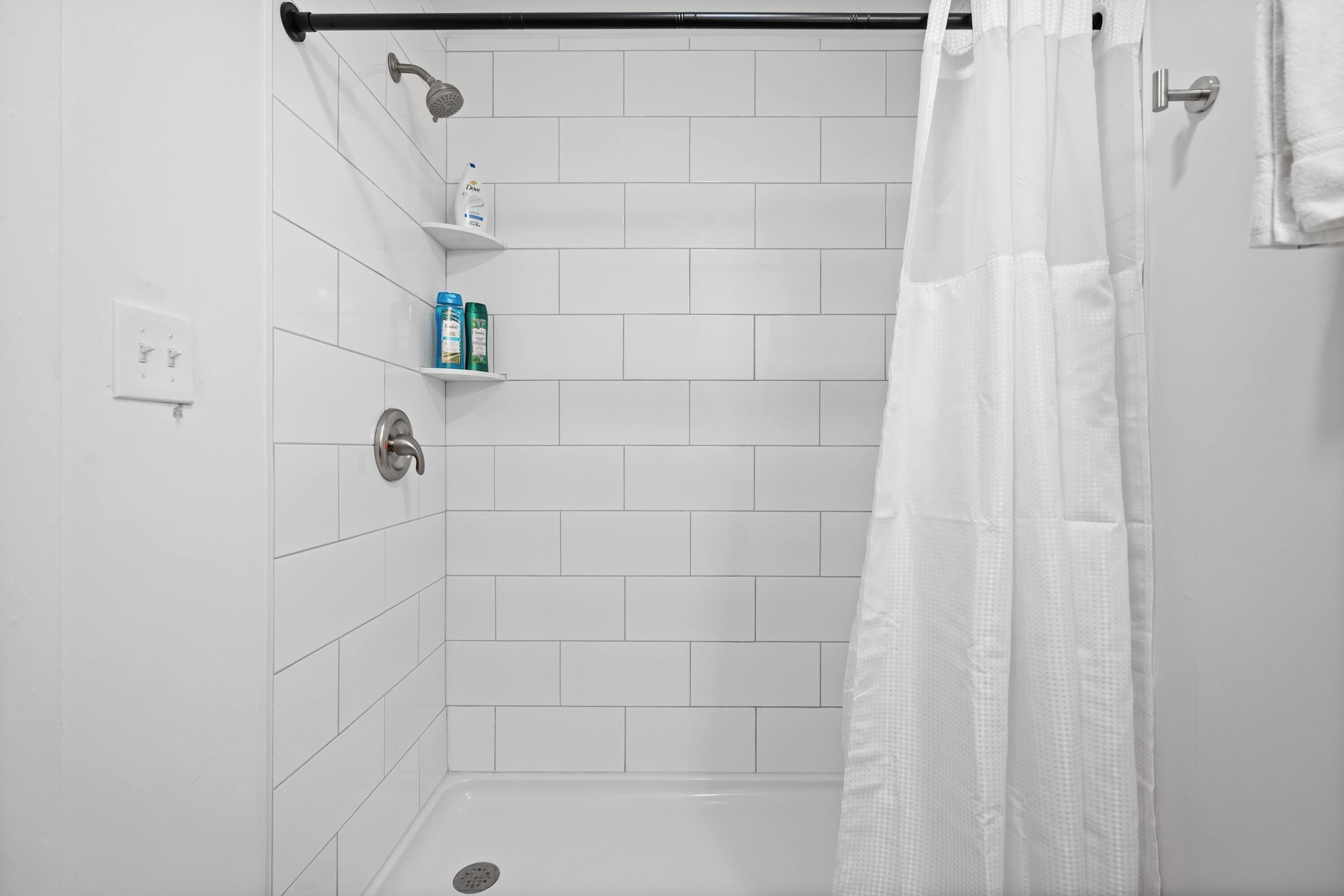 Um chuveiro com azulejos brancos, um chuveiro, uma prateleira com produtos de higiene e um cortina de chuveiro branca.