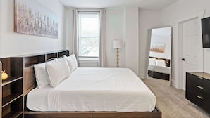 Un dormitorio con cama, mesitas de noche, ventilador de techo, ventana con cortinas, lámpara, televisor y puerta.