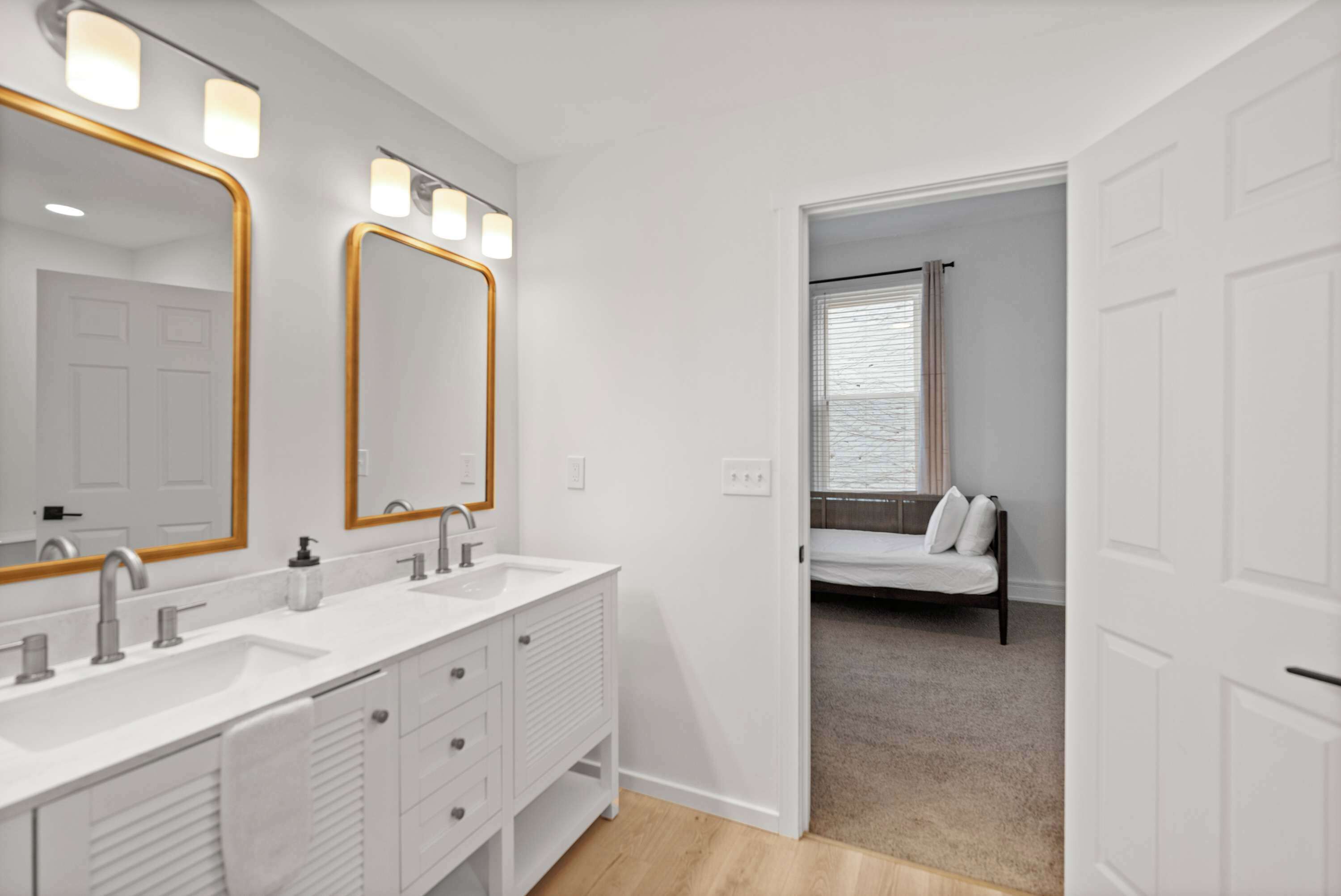 A modern bathroom with two sinks, a large mirror, and a walk-in closet leading to a bedroom with a bed and window.