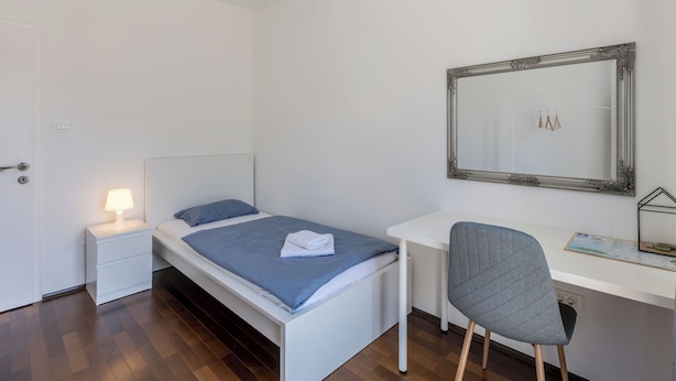 Ein Schlafzimmer mit einem Einzelbett, einem Schreibtisch, einem Stuhl, einem Nachttisch mit Lampe und einem Spiegel an der Wand.