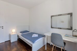 Une chambre avec un lit simple, un bureau, une chaise, une table de chevet avec une lampe et un miroir au mur.