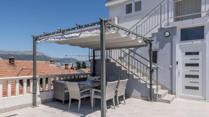 Eine Außenpatio mit Tisch und Stühlen, eine Pergola mit weißem Baldachin und eine Treppe, die zu einer oberen Etage führt.