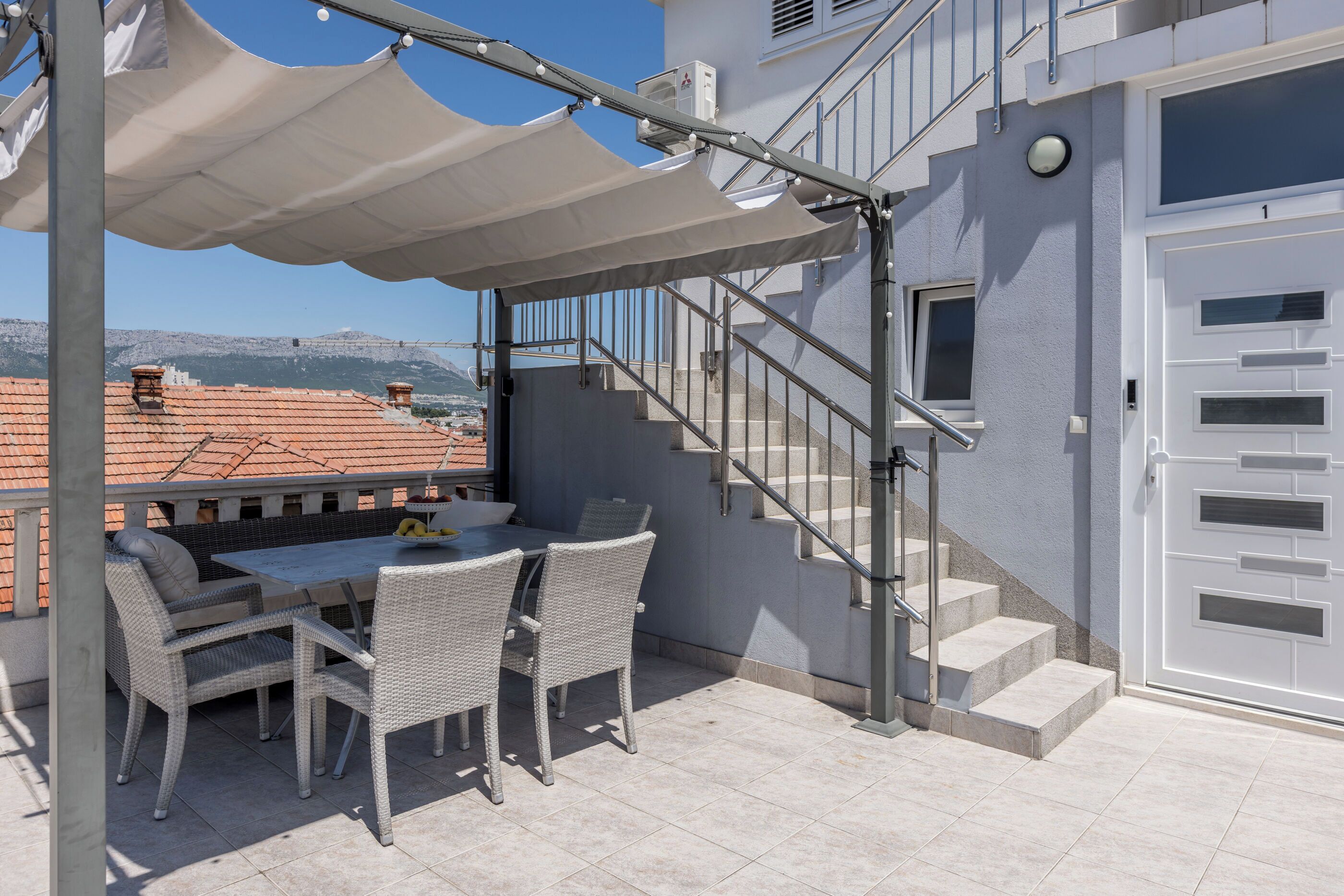 An outdoor patio with a table and chairs, a covered area, a staircase, and a view of buildings and mountains in the background.