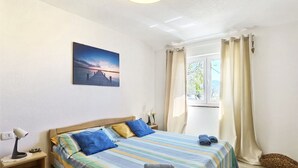 A bedroom with a bed, a nightstand, a lamp, a window with curtains, and a painting on the wall. - Apartments Adria (Karlobag)