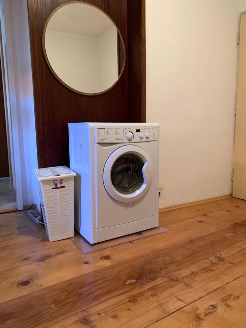 A compact, white front-loading washing machine in a room with wooden flooring and a wooden door.