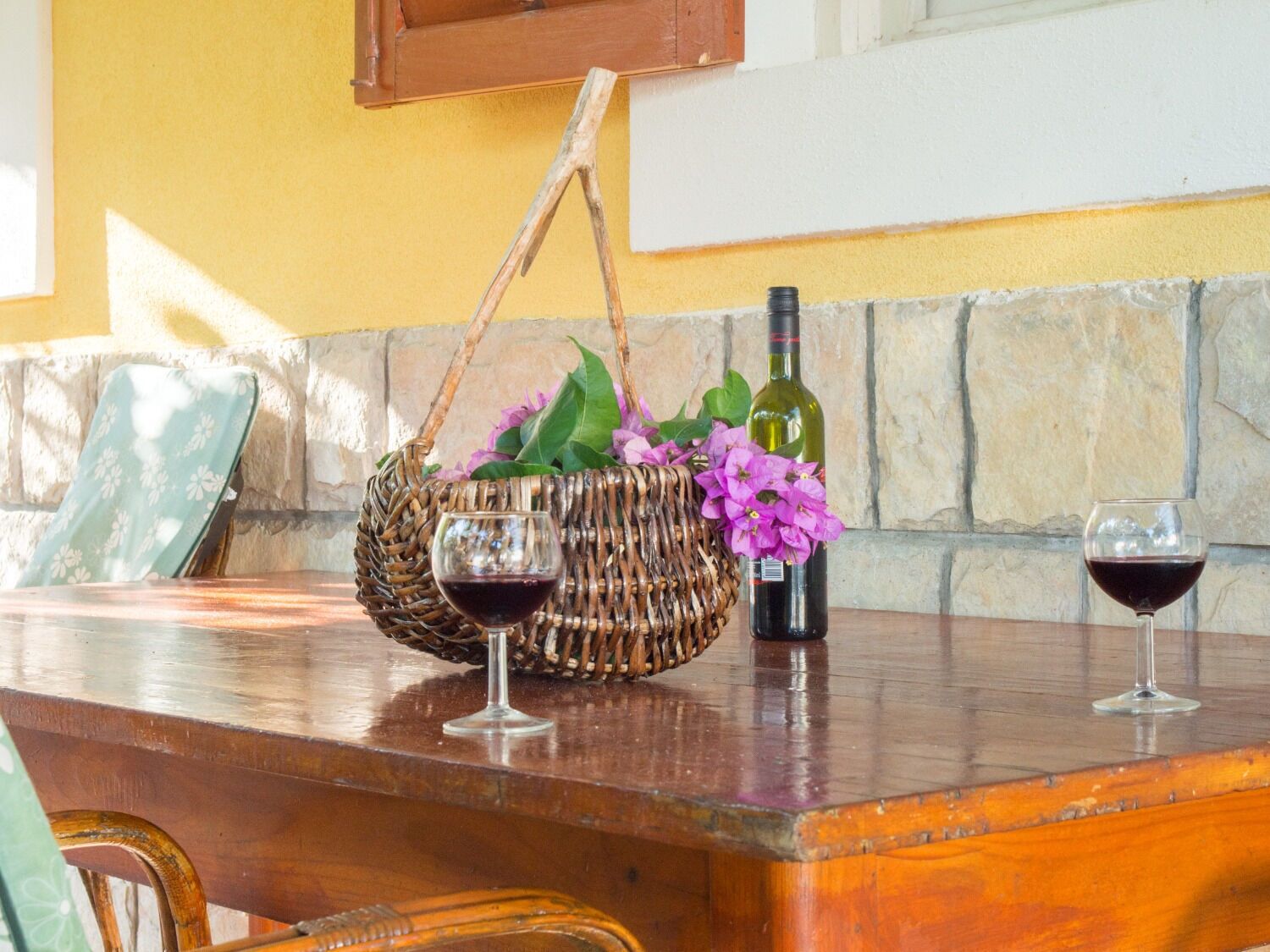 Una mesa de madera con una cesta de mimbre que contiene flores moradas y una botella de vino. Dos copas de vino están sobre la mesa.