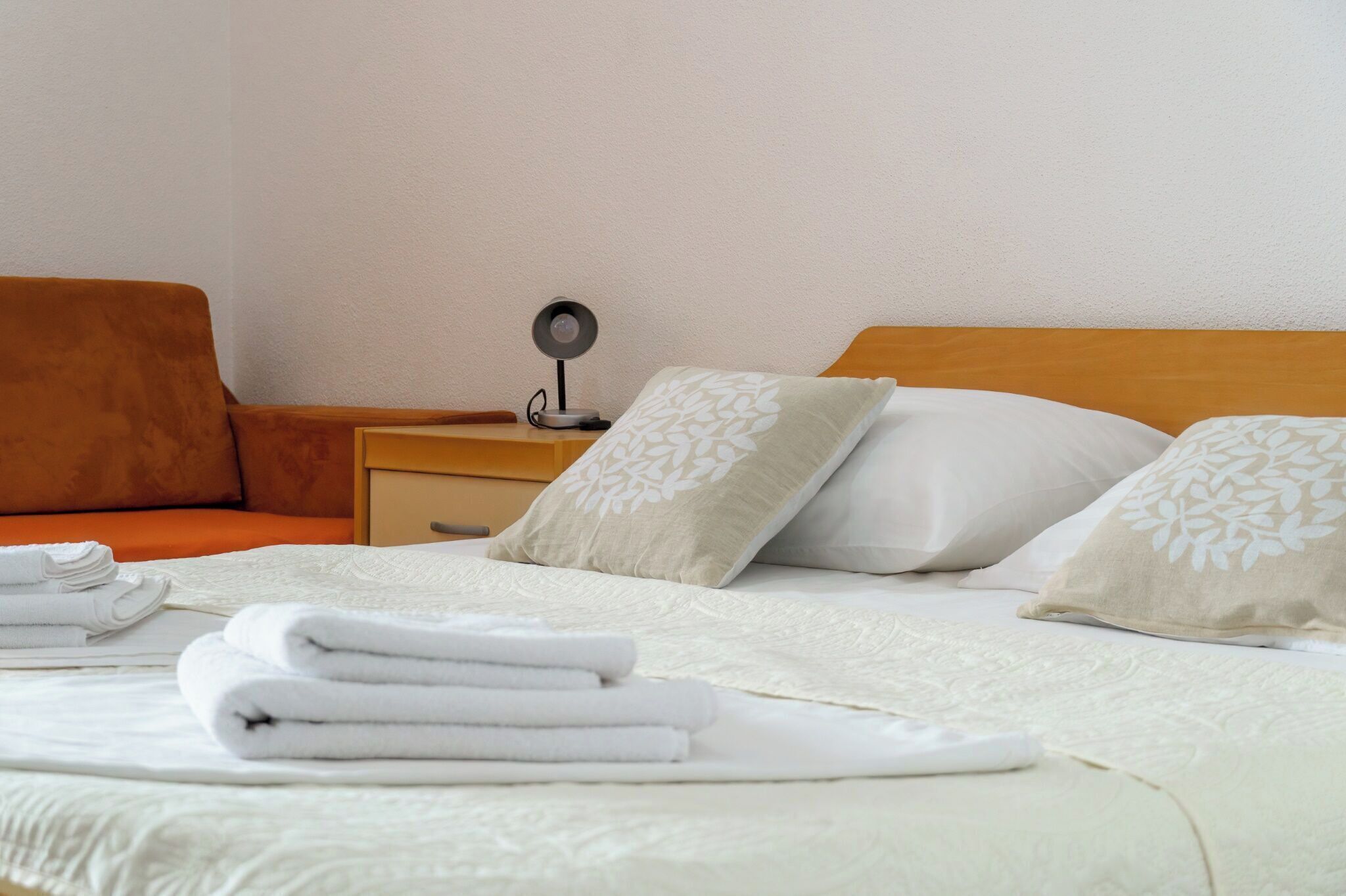 Ein ordentlich bezogenes Bett mit weißen Leinen und Kissen, ein Nachttisch mit Lampe sowie ein orangefarbener Sitzsessel.