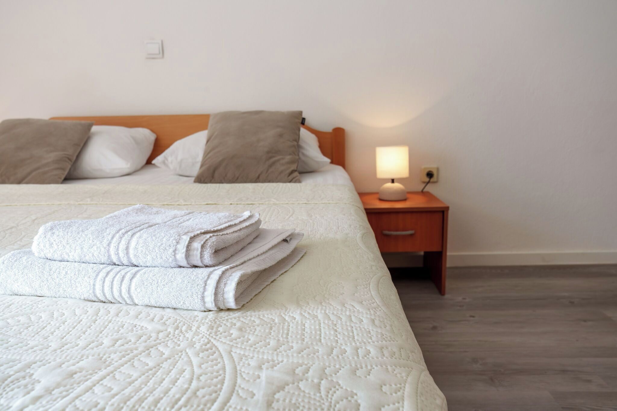 Cama bem arrumada com colcha branca e toalhas dobradas, cabeceira de madeira e mesa de cabeceira com abajur.