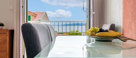 En balkong med ett glasbord och en stol, en tallrik med frukt och utsikt över en byggnad med ett rött tak.