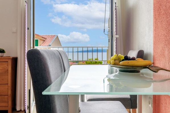 A balcony with a glass table and a chair, a plate of fruit, and a view of a building with a red roof.