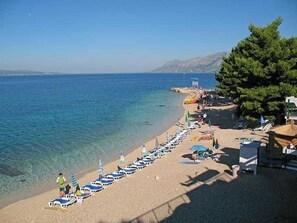 A beach with rows of sun loungers and umbrellas, a clear blue sea, and a mountainous backdrop. - Apartments "ruza" Baska Voda (Baska Voda)