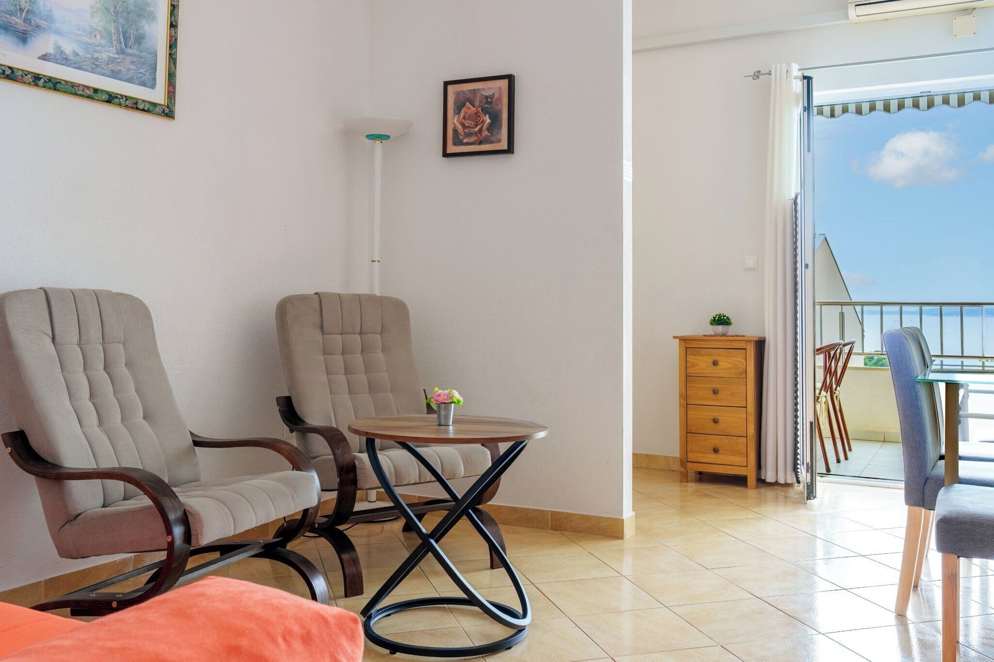 A living room with two armchairs, a small round table with a flower, a tall floor lamp, a wooden cabinet, and a balcony with a railing.