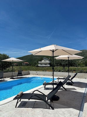A swimming pool with lounge chairs and umbrellas, surrounded by a concrete deck and a hillside in the background. - House Cord Dugopolje (Dugopolje)