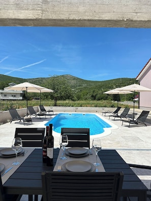 Ein Essbereich am Pool mit einem Tisch für zwei Personen, Liegestühlen und Sonnenschirmen.
