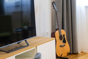 Una guitarra apoyada en una pared, un televisor sobre un soporte de madera y una ventana con cortinas.