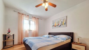 A bedroom with a bed, a nightstand, a lamp, a ceiling fan, a window with curtains, and a painting on the wall. - Apartment Emma (Novi Vinodolski)