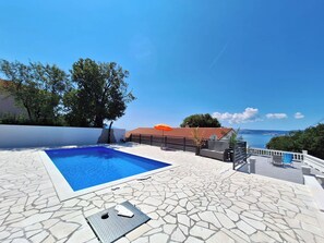 A swimming pool with a stone-tiled deck, a view of the sea, and a building with a red-tiled roof. - Apartment Emma (Novi Vinodolski)