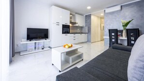 Una sala de estar moderna con un televisor de pantalla plana, una mesa de centro blanca con fruta, un sofá gris y una zona de cocina con armarios blancos y un horno empotrado.