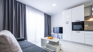 Una sala de estar moderna con un sofá, una mesa de centro con fruta, un televisor y una cocina con armarios y electrodomésticos.