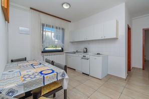 A compact kitchen with white cabinets, a dining table with a patterned tablecloth, and a window with sheer curtains. - Apartments Tomy (Medulin)
