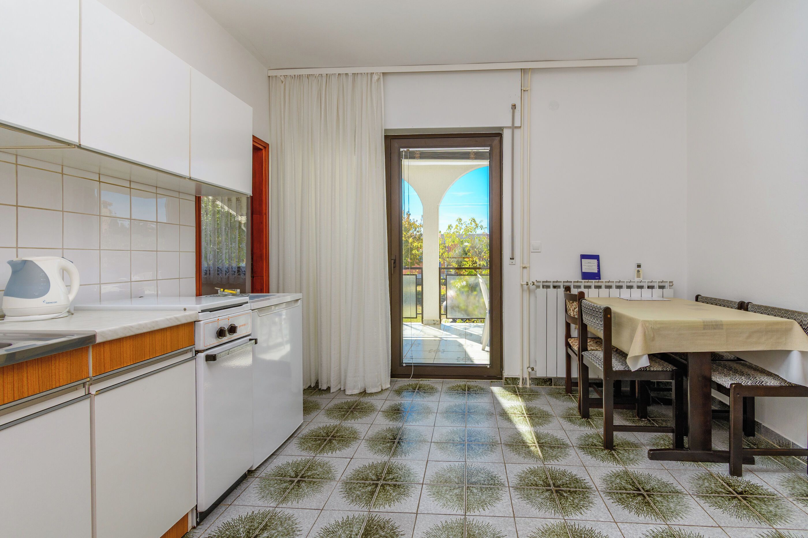 Cocina compacta con armarios blancos, estufa y pileta. Hay un comedor con mesa y sillas, y una vista a un patio exterior a través de una puerta de vidrio.
