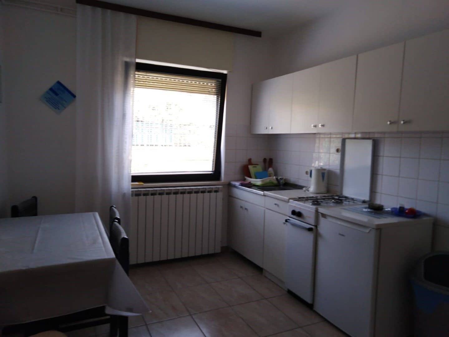 A small kitchen with white cabinets, a window with blinds, a radiator, and a table with chairs.