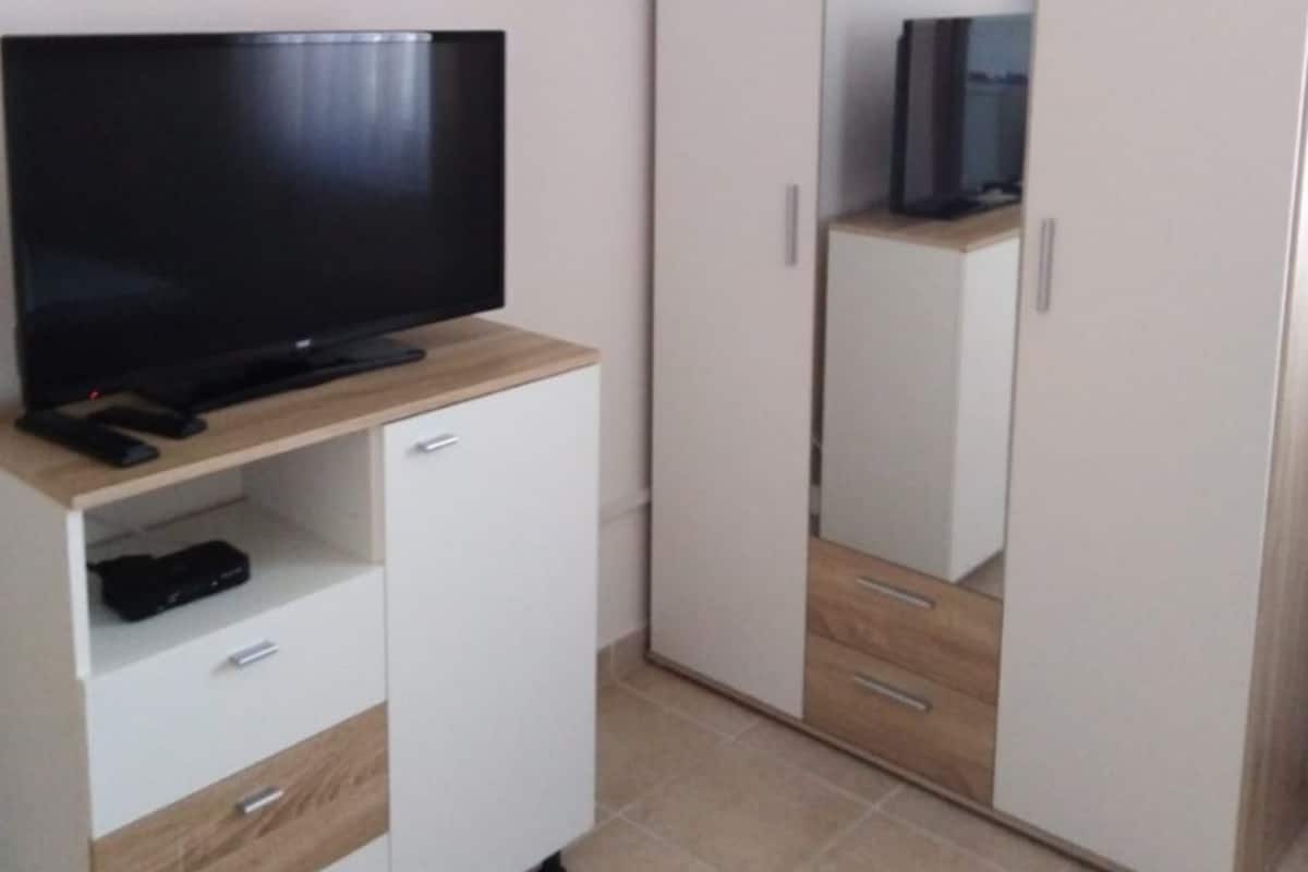 A room with a television on a wooden cabinet, a wardrobe with mirrored doors, and an air conditioning unit on the wall.