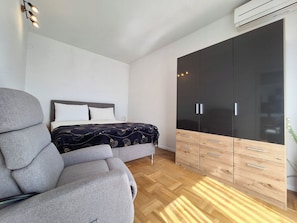 Ein modernes Schlafzimmer mit grauem Sofa, einem Bett mit gemustertem Überwurf, einem großen Kleiderschrank mit vielen Schubladen und einer Wandklimaanlage.