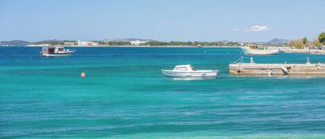 A coastal scene with boats docked at a pier, clear turquoise water, and a distant shoreline with buildings and greenery.