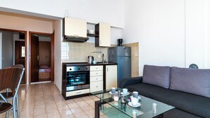 A compact kitchen with a built-in oven, a coffee machine, and a refrigerator. There is a glass dining table with cups and a chair. A sofa is placed in the living area. - Apartments Brodarica Beach (Sibenik)