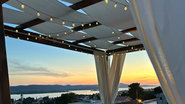 Eine Dachterrasse mit Blick auf einen Sonnenuntergang über dem Wasser, ein Tisch mit Kerze und eine überdachte Sitzgelegenheit.