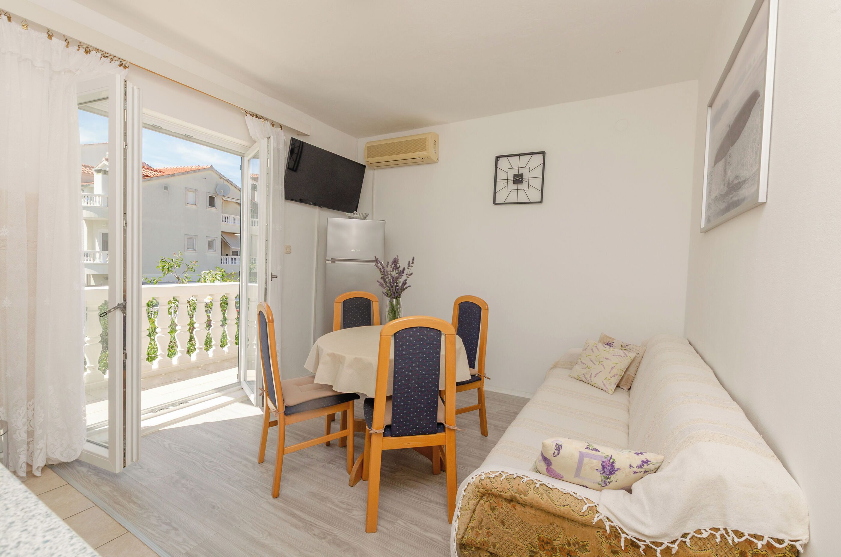A room with a balcony, a dining table with chairs, a sofa, a TV mounted on the wall, and an air conditioning unit.