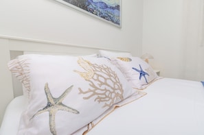 A bed with white bedding and starfish and coral patterns, a painting above the bed, and a bedside table with a lamp. - Apartments Big (Vodice)