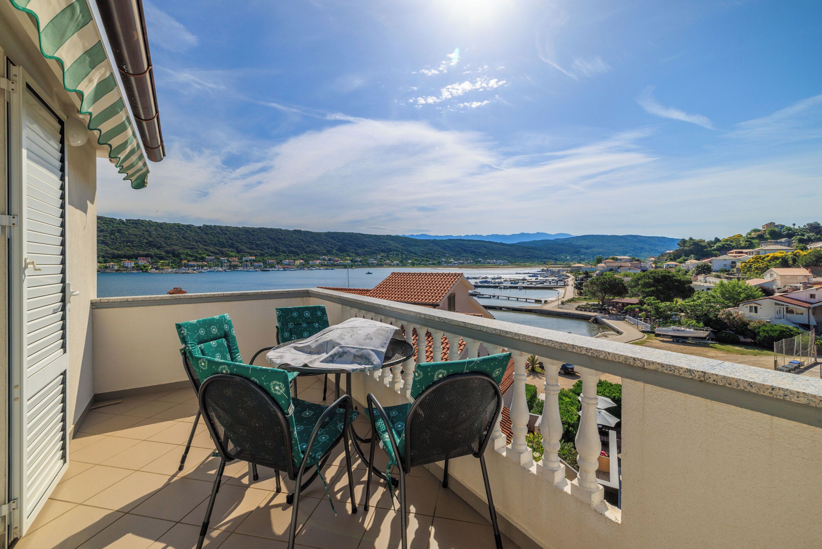 A balcony with a table and chairs, overlooking a body of water and a hilly landscape.