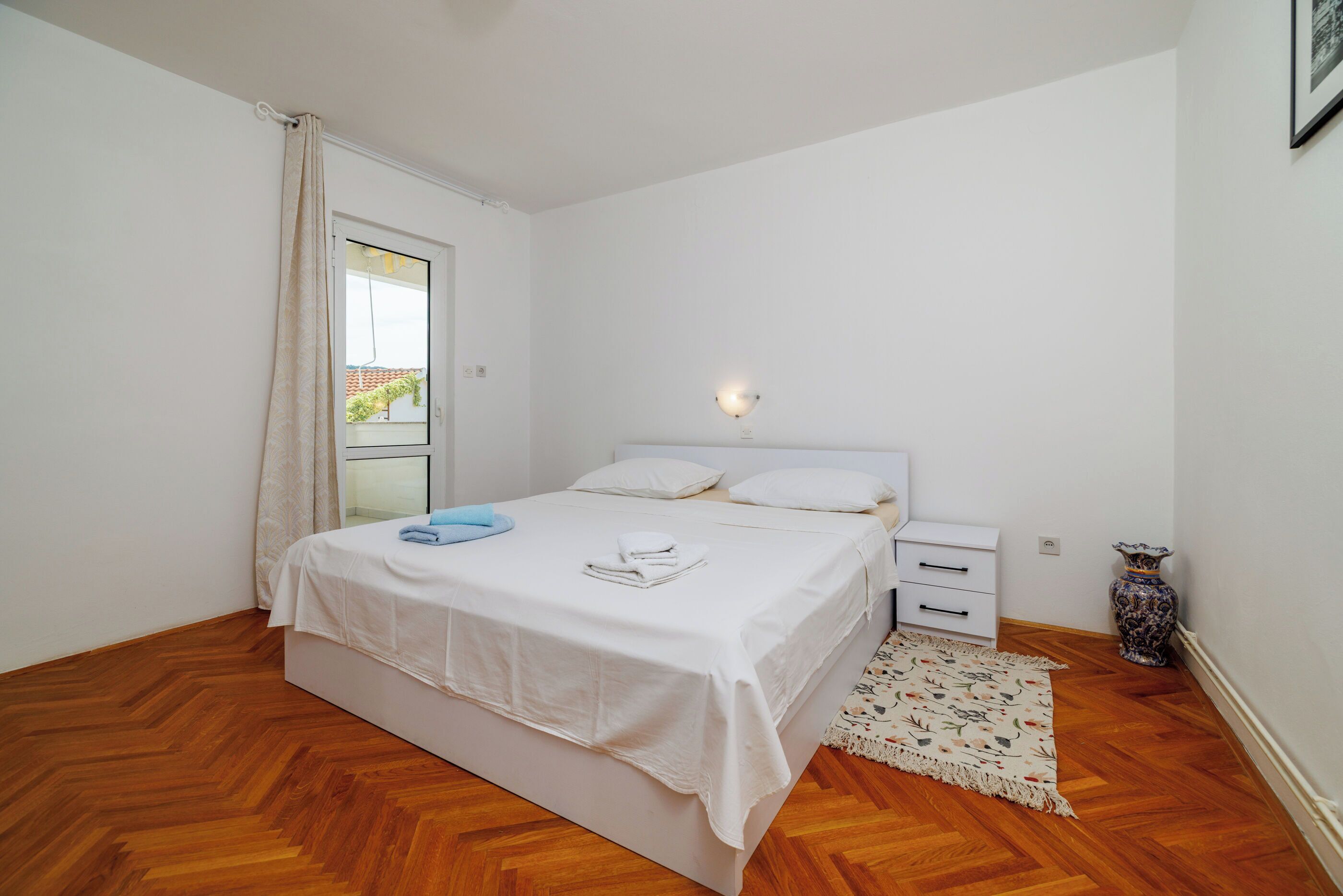 Un dormitorio con cama, mesita de noche, lámpara, ventana con cortinas y un jarrón decorativo.