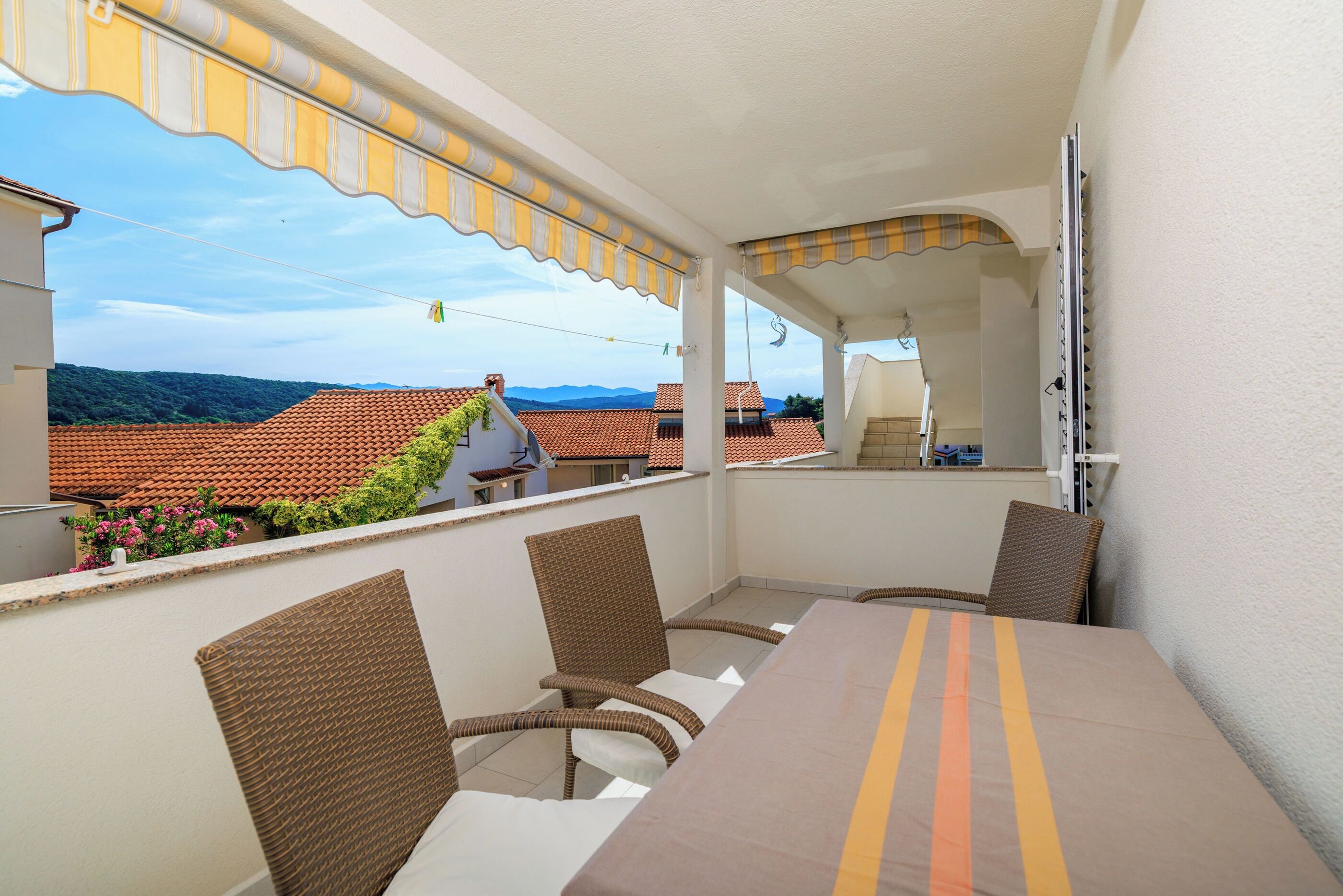 A balcony with wicker chairs and a table, overlooking a cityscape with terracotta roofs.