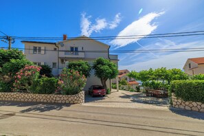 A two-story house with a car parked in the driveway, surrounded by greenery and a stone wall. - Apartments Tariba (Rab)