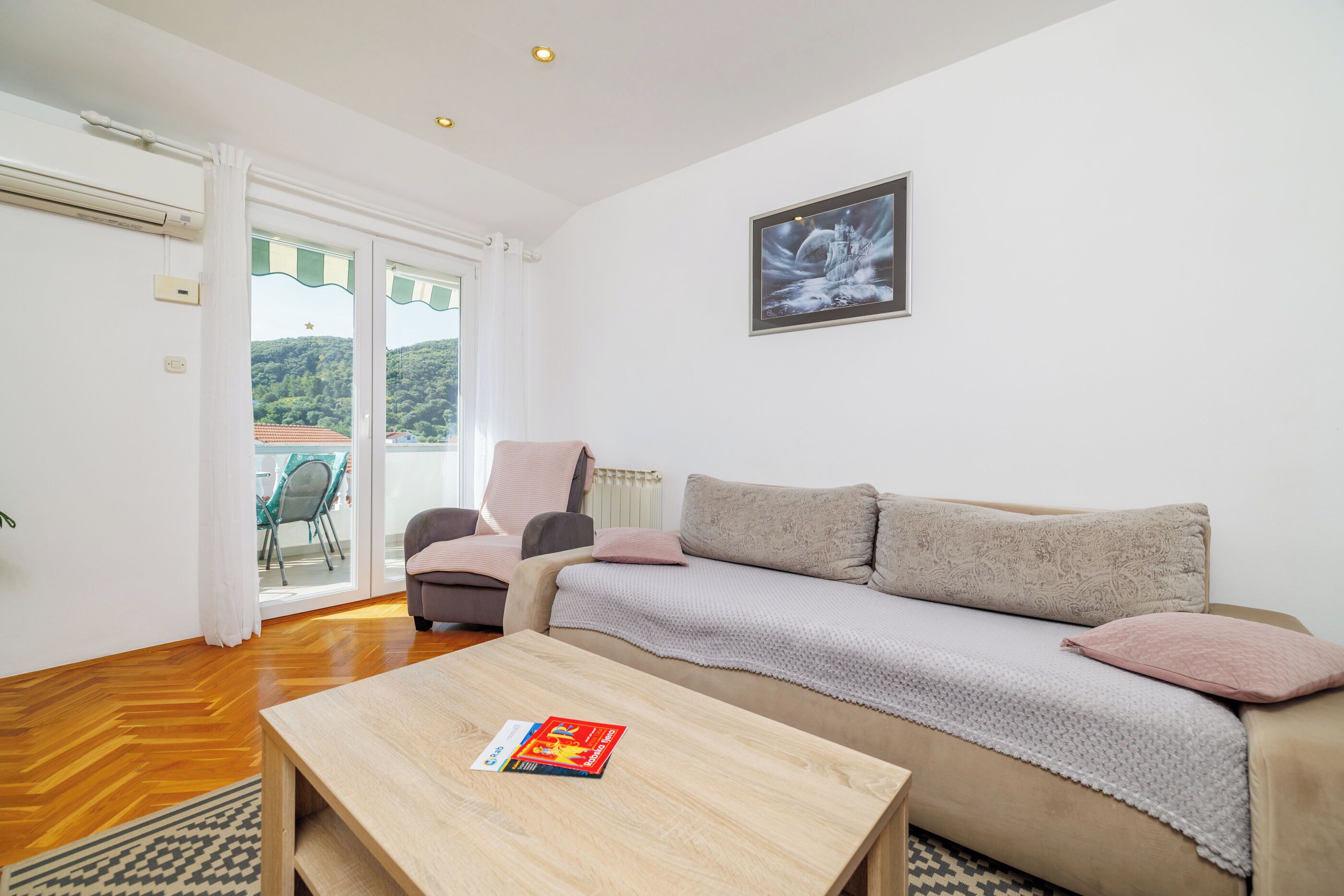 A living room with a sofa, a coffee table, and a chair. There is a window with a view of greenery and a building.