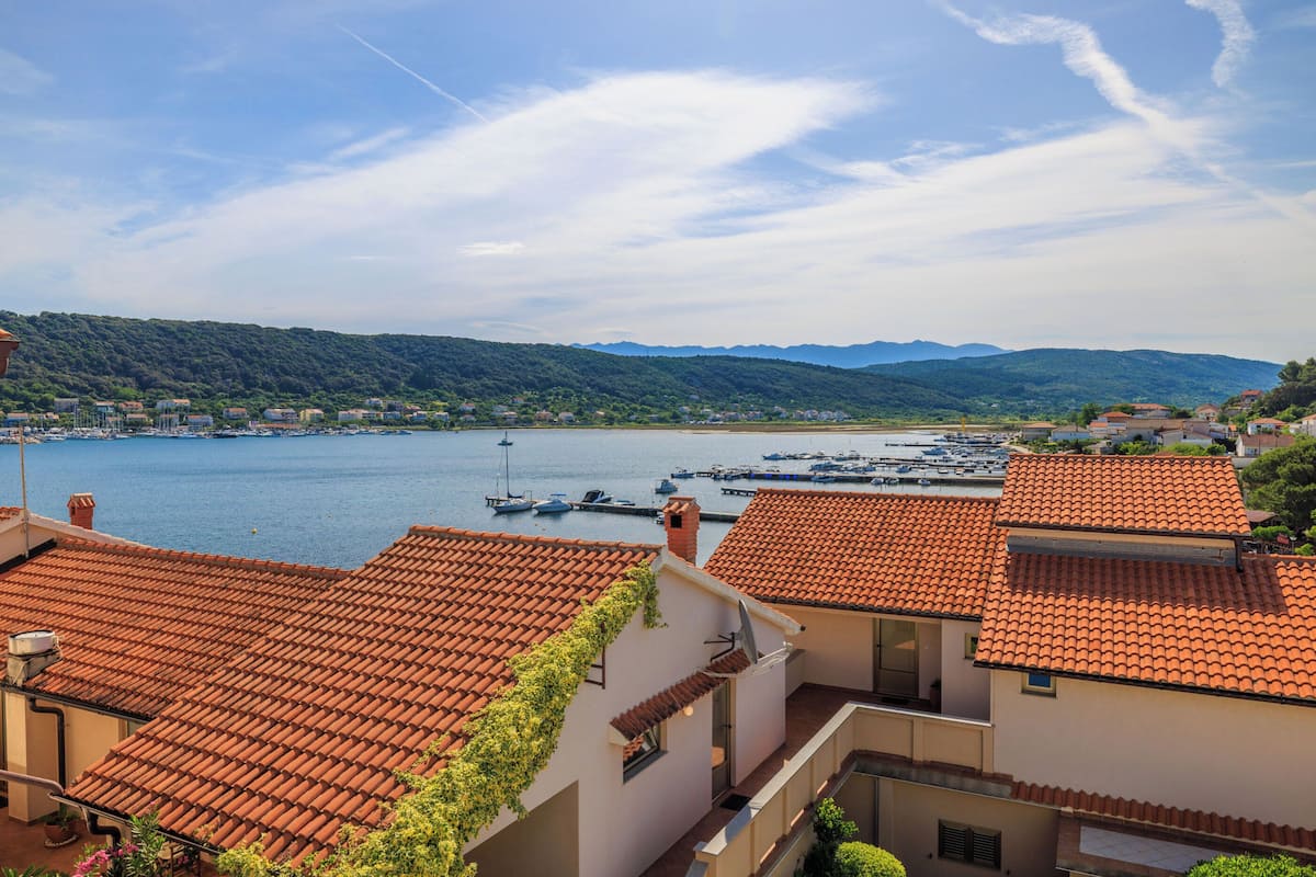 A coastal town with terracotta-roofed buildings, a marina with boats, and a backdrop of green hills.