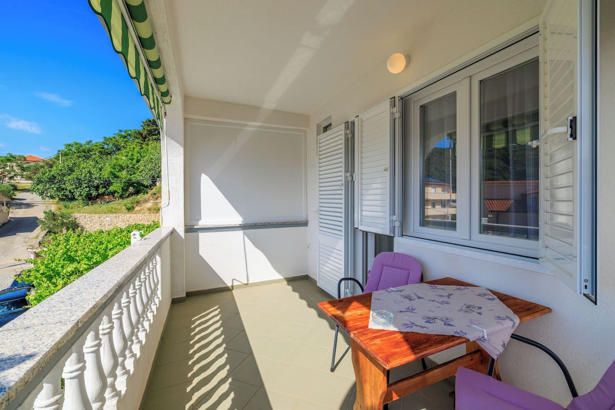 A balcony with a wooden table and chairs, a window with shutters, and a view of greenery and a road.