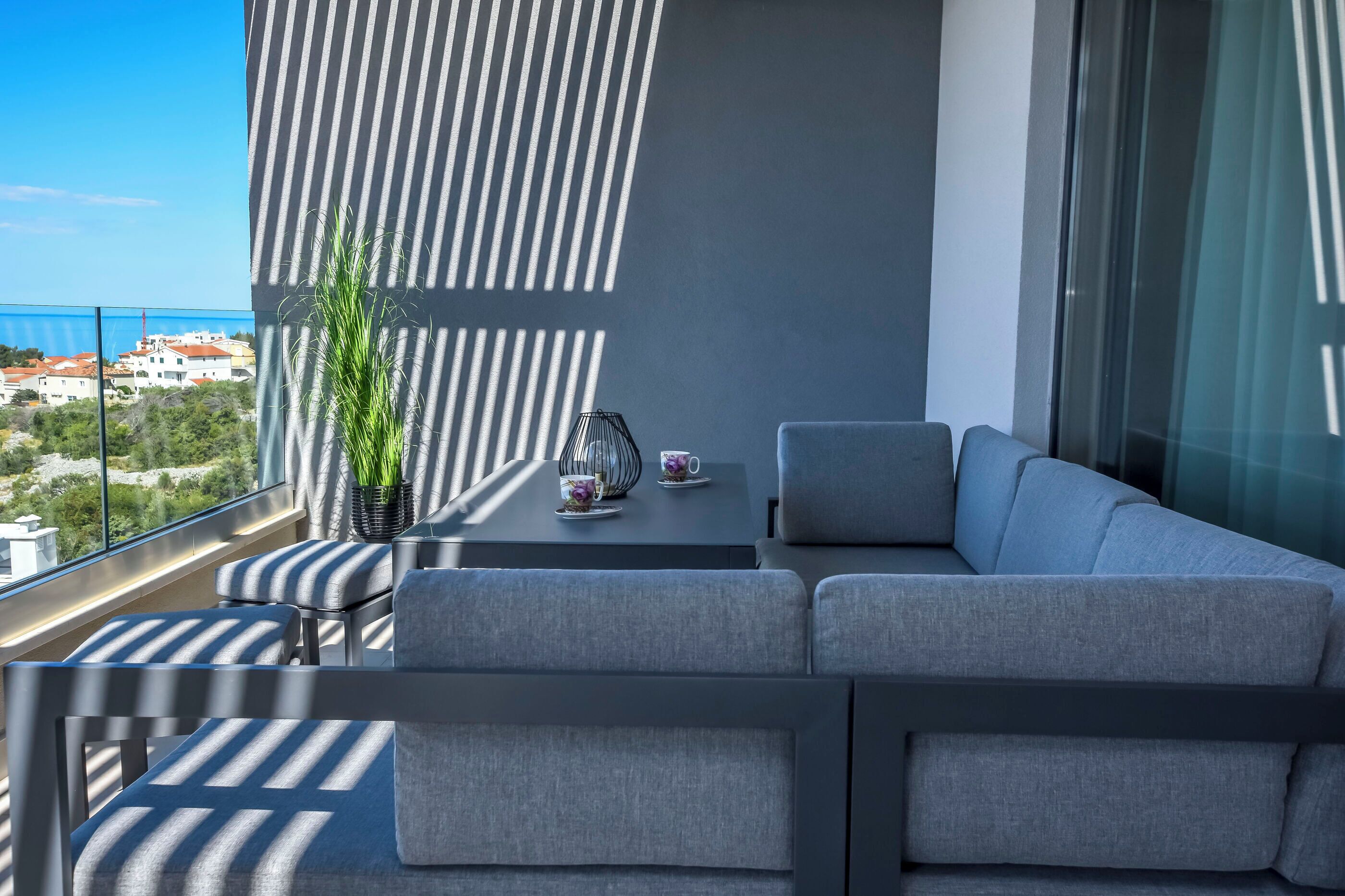 A modern balcony with a grey sofa, a small table with a potted plant and a teapot, and a view of the cityscape.