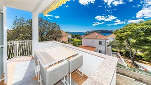 Un balcón con una mesa blanca y sillas de mimbre, con vista a un pueblo costero con casas y árboles.