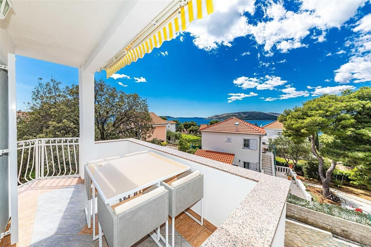 Ein Balkon mit weißem Tisch und Korbstühlen, mit Blick auf ein Küstendorf mit Häusern und Bäumen.